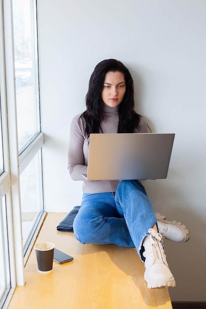 A woman sits on a window sill and looks at a laptop.