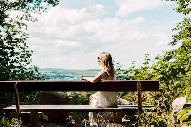 Женщина сидит спиной на скамейке и смотрит на город и горы