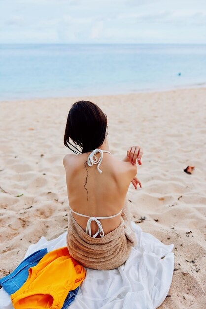 A woman sits with her back to the camera after swimming in the ocean in a swimsuit on a towel on the sand and looks at the sea and clouds summer beach vacation