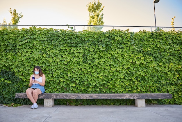 A woman sitting alone on a bench with a virus mask.