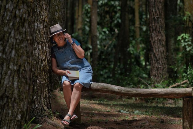 Photo woman sitting alone on log bench in shady forest making a phone call using smartphone