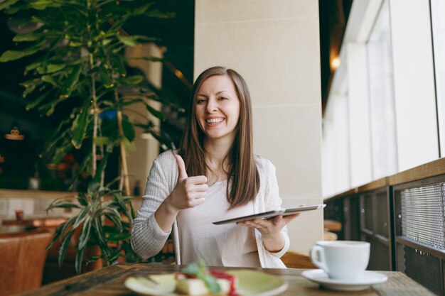 Woman sitting alone near big window in coffee shop at table with cup of cappuccino, cake, relaxing in restaurant during free time. Female working on pc tablet computer rest in cafe. Lifestyle concept.