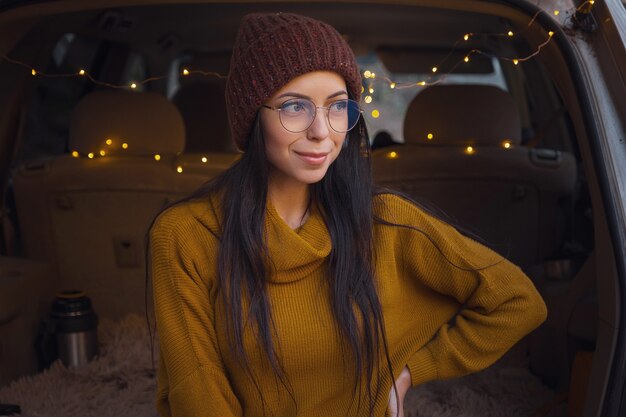 Woman sitting in back of car trunk. Getting ready to go. Young laughing woman sitting in the open trunk of her auto. Autumn road trip. Hipster toad travel, Fall Getaway