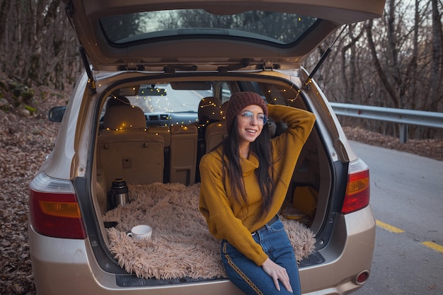 車のトランクの後ろに座っている女性。行く準備をしています。彼女の自動車の開いたトランクに座っている若い笑う女性。秋のロードトリップ。流行に敏感なヒキガエルの旅行、秋の逃走