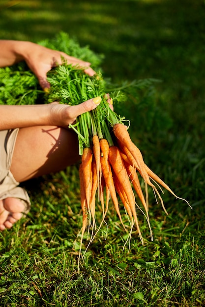 Woman sitting barefoot on grass with branch of raw organic carrots