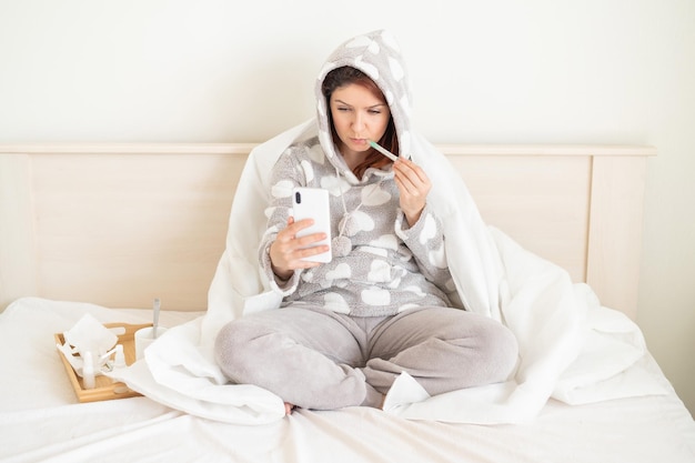 Woman sitting on the bed covered with a blanket and photographed on the phone with a thermometer in his mouth A girl with a cold takes a selfie on her blog Measurement of elevated temperature