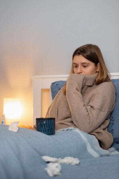 ベッドに座って気分が悪い女性 若い女の子は温かい飲み物を飲み、病気の治療法をとります