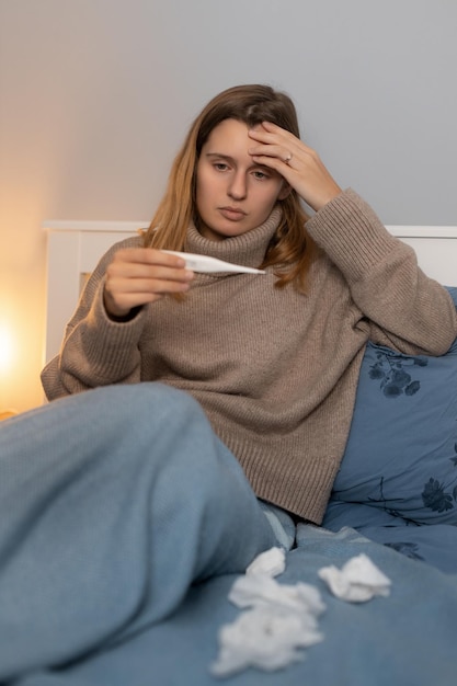 Woman sitting in bed touching head and looking on thermometer\
girl measures body temperature