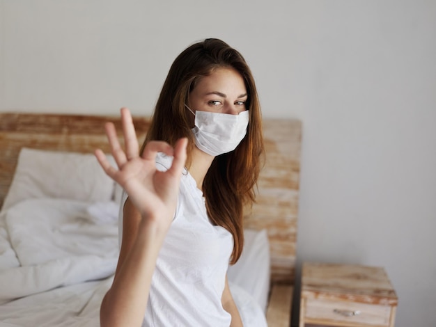 Woman sitting on bed wearing medical mask positive hand gesture