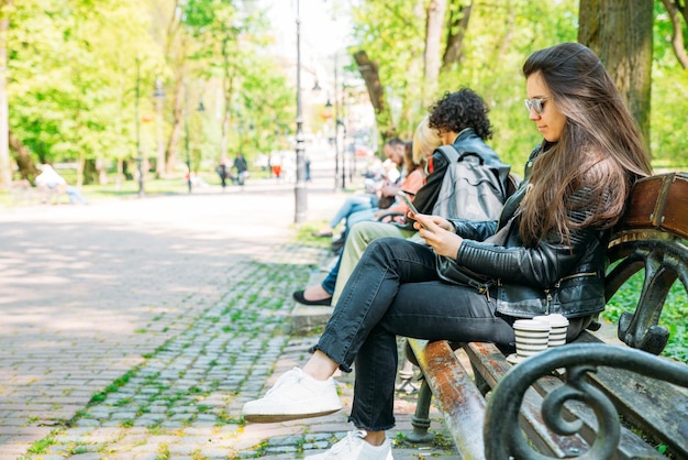 Woman sitting on bench in city park drinking coffee surfing internet via mobile