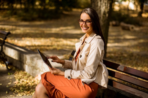 タブレットPCを使用して、ソーシャルメディアをチェックして秋の天候の間に公園のベンチに座っている女性。