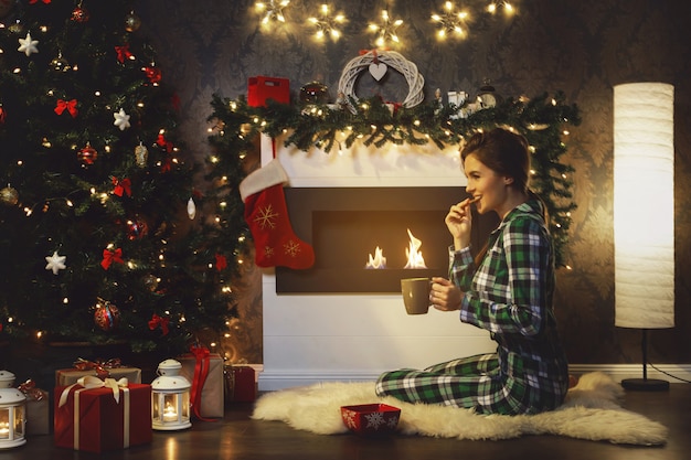 Woman sitting beside fireplace is drinking hot tea with cookies