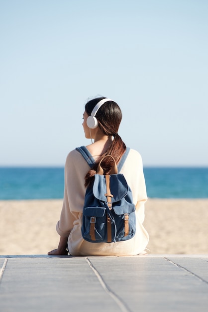 Женщина, сидящая на море с наушниками и рюкзаком
