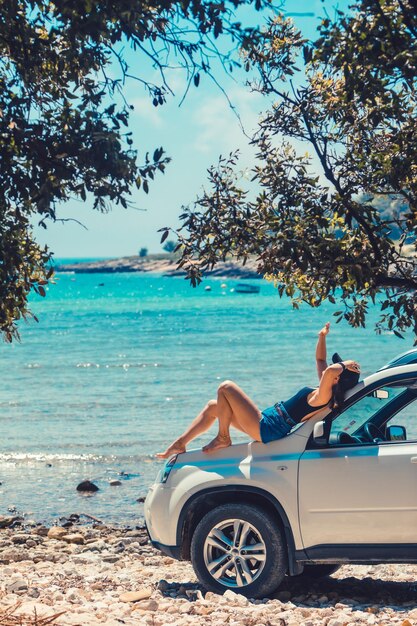 海のビーチの海辺を見ている車のフードに座っている女性