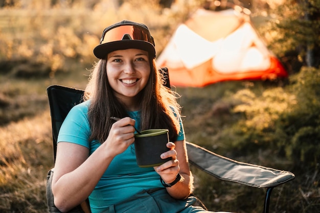 Woman sitting in chairs outside the tent sunset camping in
forest recreation outdoor activity cooking dinner with camping gear
in camp summer travel outdoor adventure