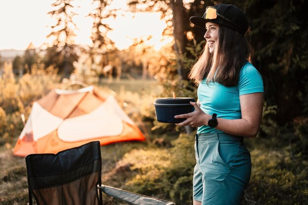 Woman sitting in chairs outside the tent sunset camping in\
forest recreation outdoor activity cooking dinner with camping gear\
in camp summer travel outdoor adventure