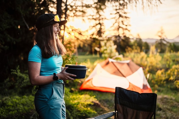 Woman sitting in chairs outside the tent sunset camping in
forest recreation outdoor activity cooking dinner with camping gear
in camp summer travel outdoor adventure