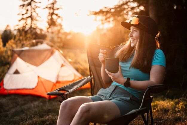 Woman sitting in chairs outside the tent sunset camping in\
forest recreation outdoor activity cooking dinner with camping gear\
in camp summer travel outdoor adventure