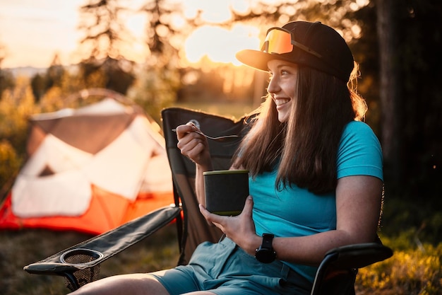 Woman sitting in chairs outside the tent sunset camping in\
forest recreation outdoor activity cooking dinner with camping gear\
in camp summer travel outdoor adventure