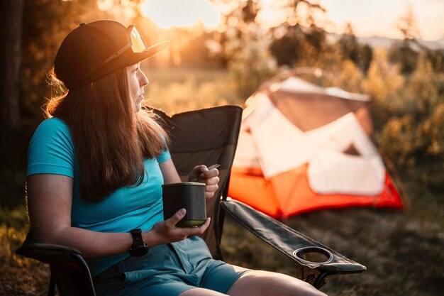 Woman sitting in chairs outside the tent sunset camping in
forest recreation outdoor activity cooking dinner with camping gear
in camp summer travel outdoor adventure