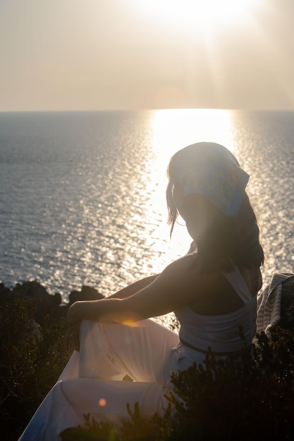 Женщина сидит у скалы и смотрит на закат над морем