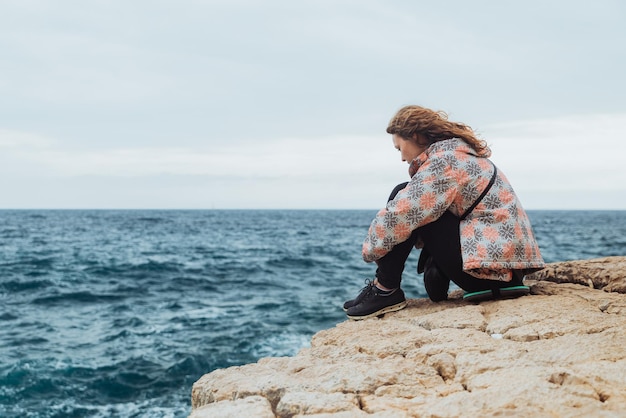 Женщина сидит на скале с грустным видом и смотрит на бушующее море