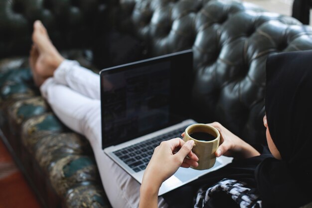 Woman sitting on couch holding cup of coffee looking at laptop