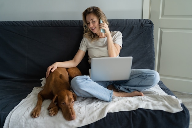 Woman sitting at couch, stroking her lovely Vizsla dog, talking on cellphone, remotely working on computer