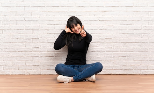 Woman sitting on the floor making phone gesture and pointing front