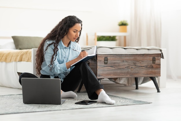 Woman sitting on floor using computer and writing in notebook