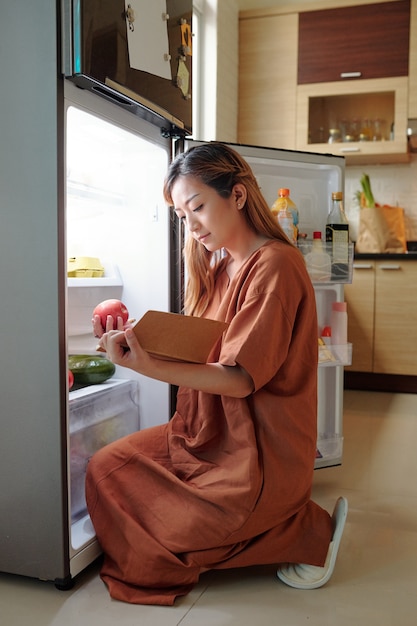 Foto donna seduta davanti al frigorifero aperto e controlla se ha tutti gli ingredienti per il piatto che sta per cucinare