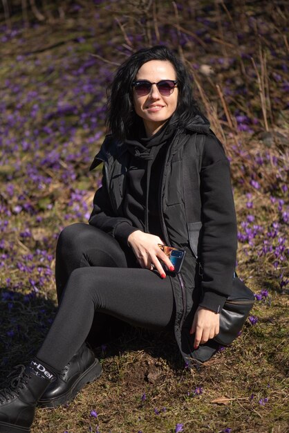 A woman sitting in the grass with her legs crossed and wearing a black jacket.