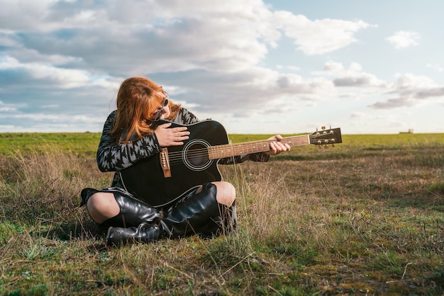 Woman sitting in a green field looking at a black guitar under a sky with clouds