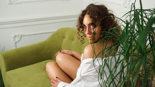 Donna seduta sul divano verde nel soggiorno. belle gambe lunghe. bella donna con capelli ricci in lingerie bianca a casa - mattina felice