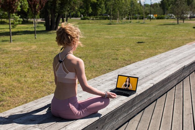 Фото Женщина, сидящая в позе йоги на открытом воздухе на деревянном променаде, смотрит фитнес-видео на ноутбуке