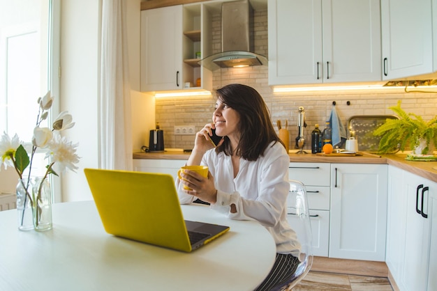 Женщина сидит на кухне, работает на ноутбуке, разговаривает по телефону и пьет чай