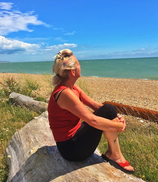 Woman sitting on log at beach against blue sky