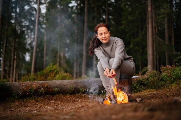 ハイキングで丸太に座っている女性が森のキャンプファイヤーで夕食のためにソーセージを調理します森の中での生存