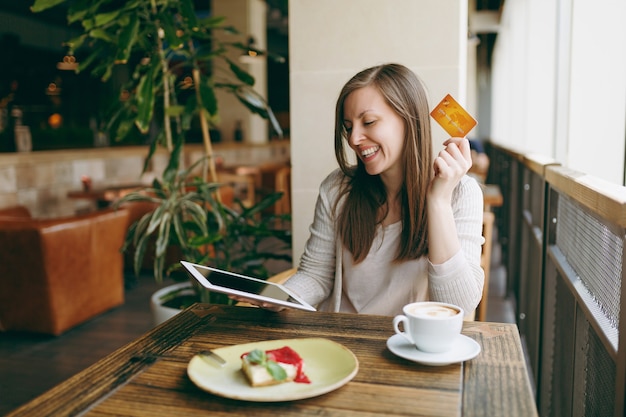 Woman sitting near big window in coffee shop at table with credit card, cup of coffee cake relaxing in restaurant during free time. Female working on tablet pc computer rest in cafe. Lifestyle concept