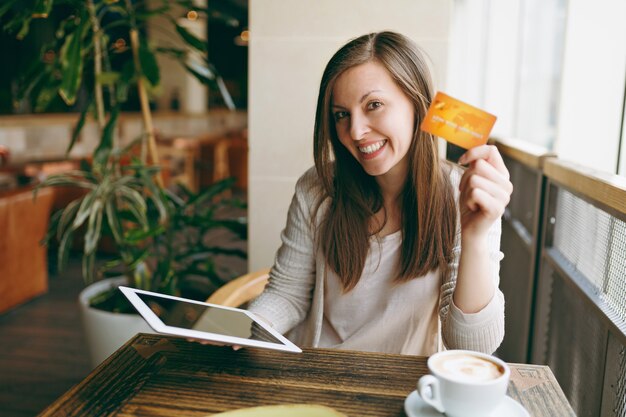 Woman sitting near big window in coffee shop at table with credit card, cup of coffee cake relaxing in restaurant during free time. Female working on tablet pc computer rest in cafe. Lifestyle concept