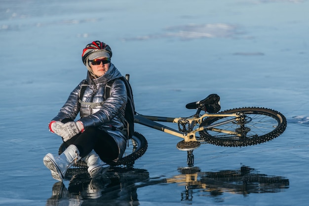 氷の上で自転車の近くに座っている女性サイクリストの女の子が休憩をとった彼女は車輪の上に座って夕日の美しい景色を楽しんでいます旅行者は自転車に乗っています女性は氷の上で自転車に乗っています