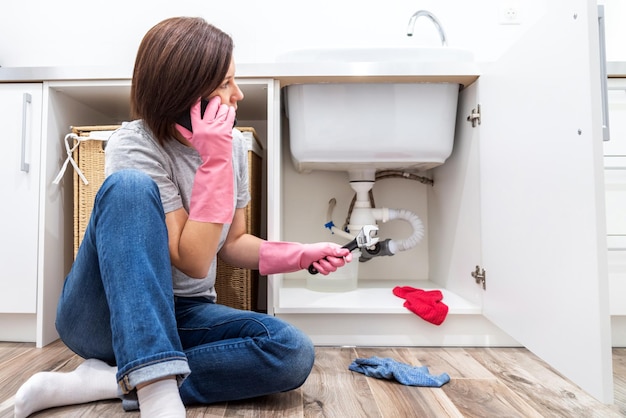 Donna che si siede vicino al lavandino colante che chiede aiuto per telefono