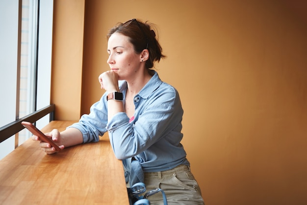 Woman sitting near window in to the cafe and using her phone. Waiting for the order