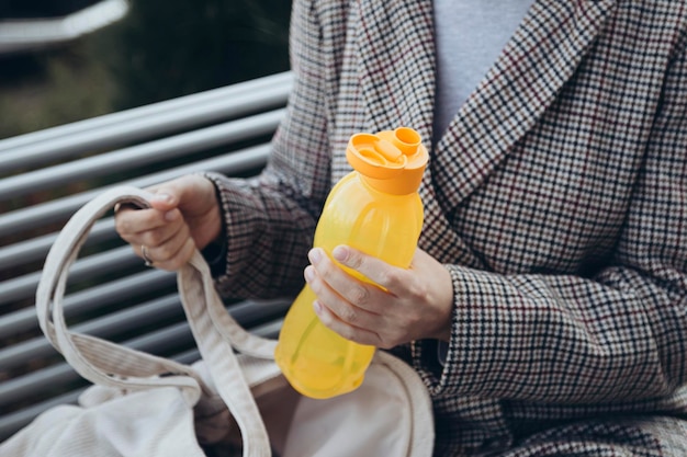 写真 手に黄色いプラスチックの水のボトルを握る屋外のベンチに座っている女性