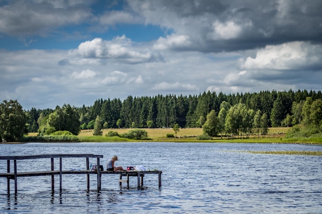 写真 雲の空に照らされた木々に囲まれた湖の上の埠頭に座っている女性