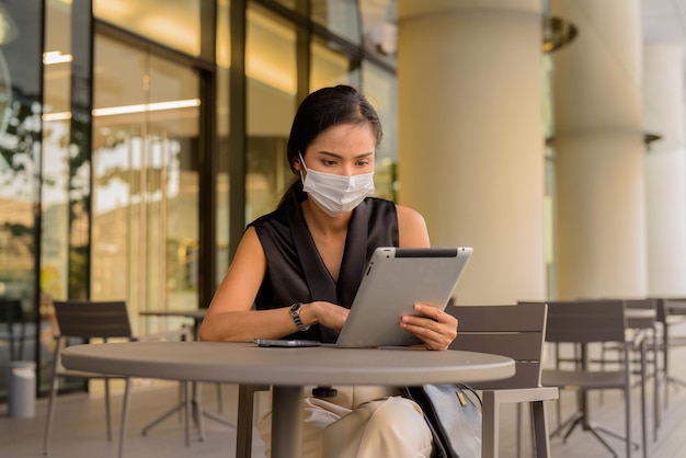 コーヒーショップのレストランで屋外に座っている女性は、電話とデジタルタブレットの水平方向のショットを使用しながら、covid19から保護するために社会的な距離とフェイスマスクを着用しています。