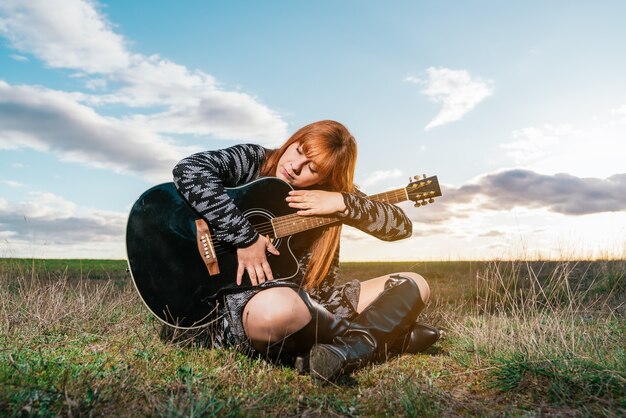 曇り空の下で彼女の黒いギターを抱き締める公園に座っている女性。音楽へのコンセプト愛