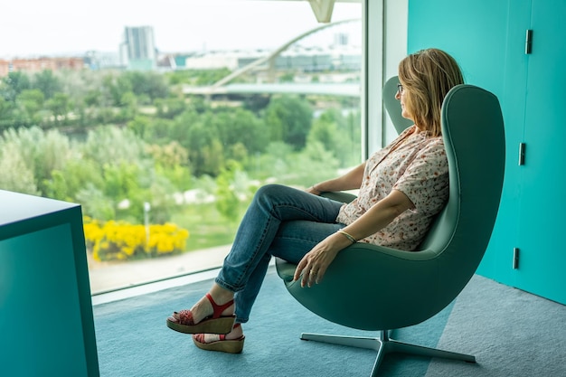 Woman sitting in a relaxed armchair and contemplating the landscape from a large window of the room