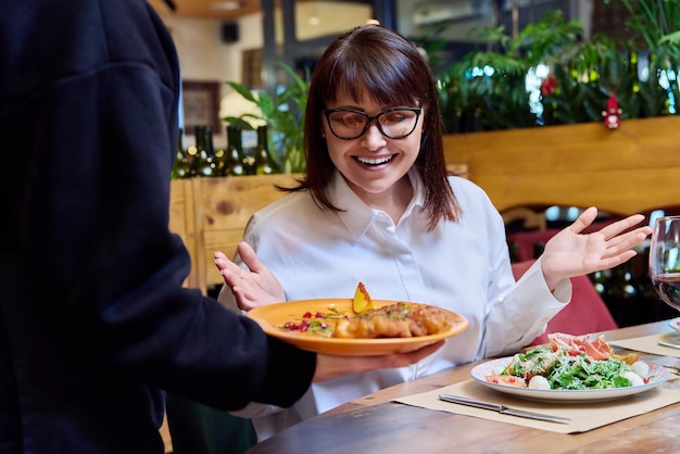 Woman sitting in restaurant rejoices at plate of cooked food in hands of waiter