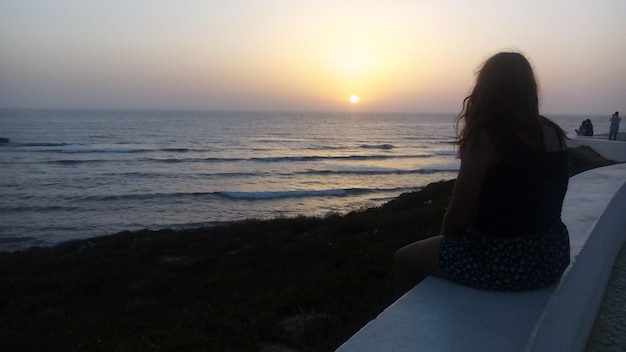 Foto donna seduta su un muro di sostegno che guarda il mare durante il tramonto